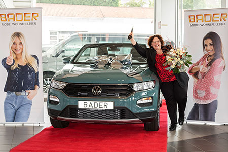 Gewinnspielübergabe vor einem mit Schleife geschmückten VW T-Roc mit BADER-Kennzeichen; eine Frau steht auf dem roten Teppich mit Blumenstrauß und Autoschlüssel.