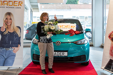 Gewinnspielübergabe vor einem mit Schleife dekorierten Elektroauto; eine Frau steht auf dem roten Teppich und hält einen Blumenstrauß.