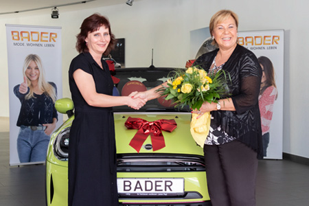 Zwei Frauen bei der Gewinnspielübergabe vor einem mit Schleife geschmückten Auto, eine überreicht einen Blumenstrauß.