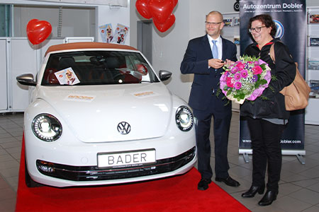 Gewinnspielübergabe vor einem weißen VW Beetle Cabriolet mit BADER-Kennzeichen; zwei Personen stehen auf dem roten Teppich, eine überreicht einen Blumenstrauß, im Hintergrund hängen rote Herzballons.